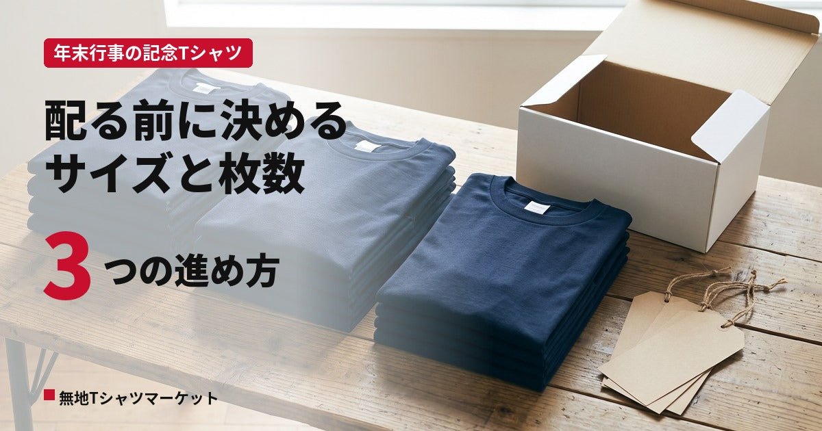 木のテーブルに三つの山へ畳み分けた濃紺の無地Tシャツと、配布用の白い箱・無地の下げ札を並べた様子